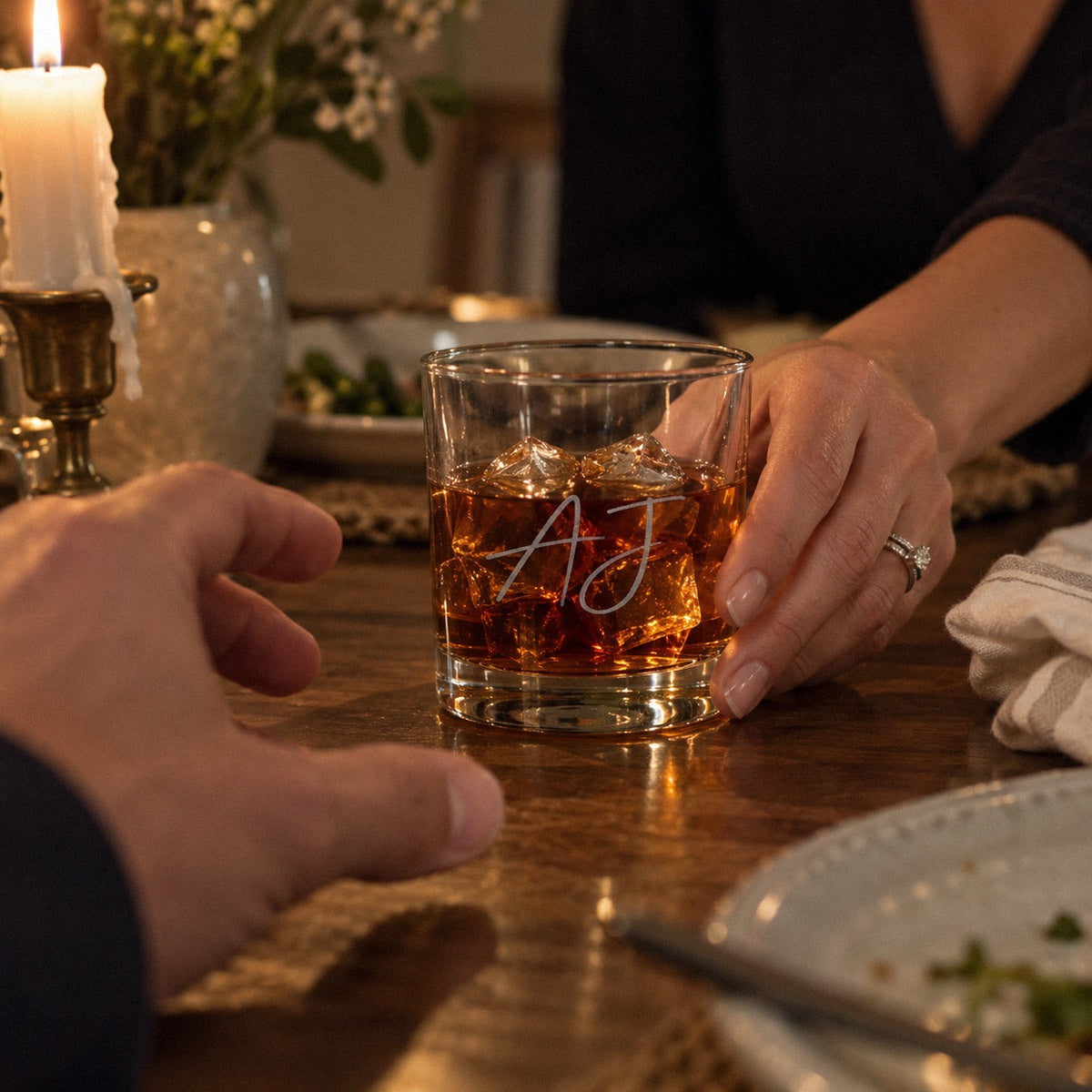 A wife slides a My Heart whiskey glass etched AJ to her husband at an anniversary dinner, a personalized Valentine's gift for him
