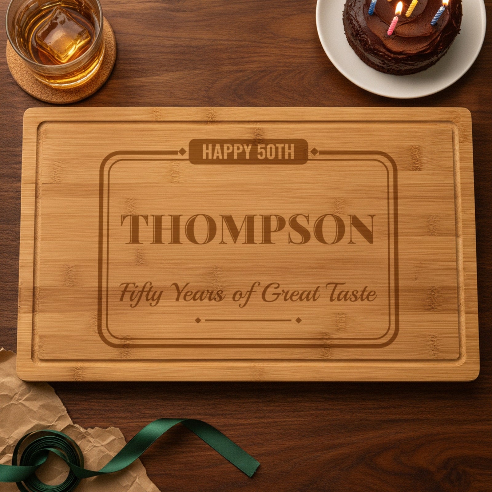 Personalized 50th birthday cutting board engraved Happy 50th, Thompson and Fifty Years of Great Taste beside a chocolate birthday cake, bourbon and unwrapped gift paper