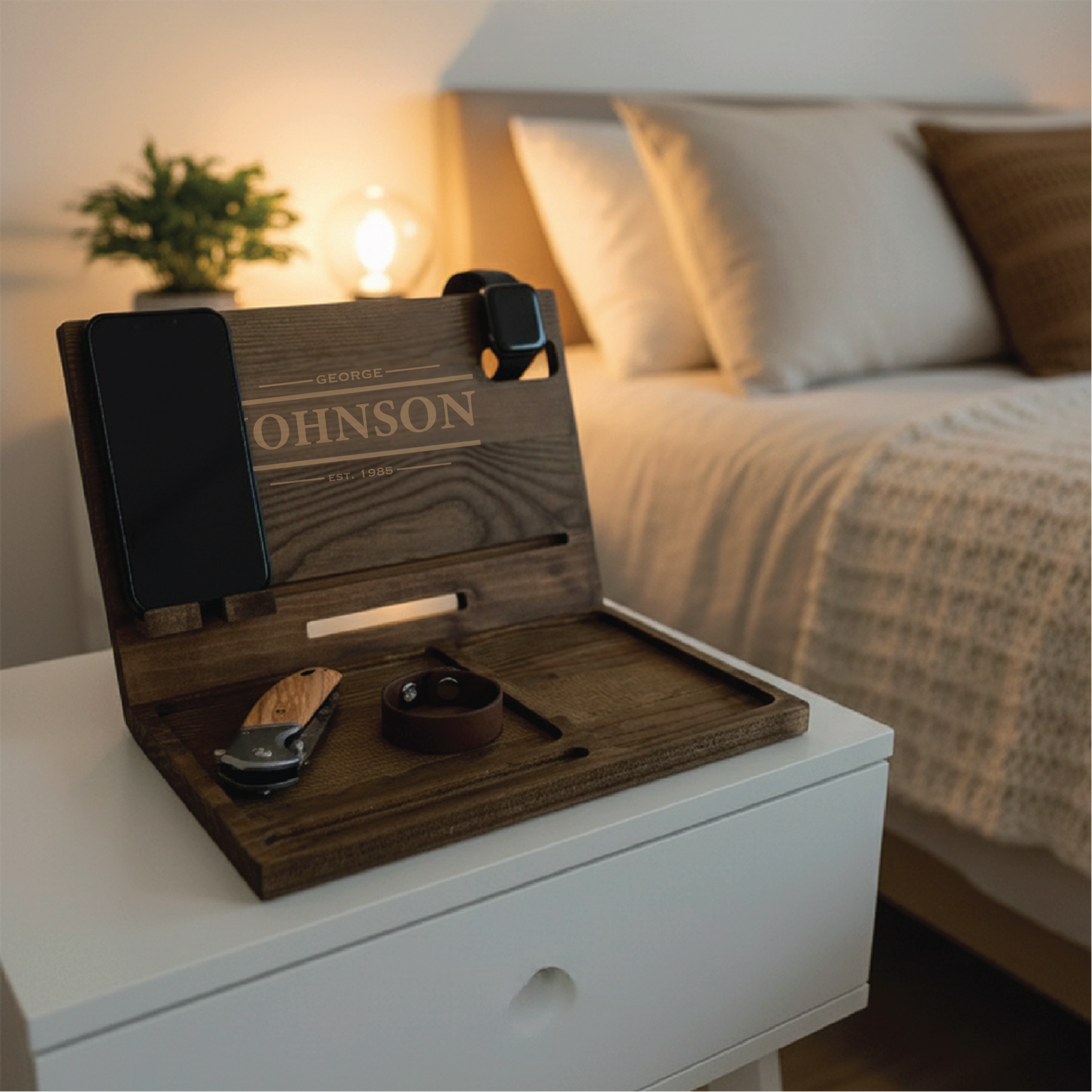 Wood desk organizer with sample engraving "GEORGE JOHNSON EST. 1985" shown in bedroom setting with phone, watch, wallet, and knife displayed in compartments