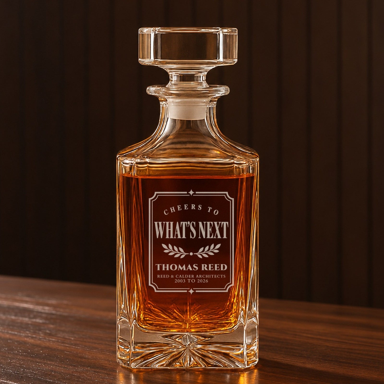 Personalized whiskey decanter with a framed Cheers to What's Next label engraved for Thomas Reed and his architecture firm, on a walnut bar