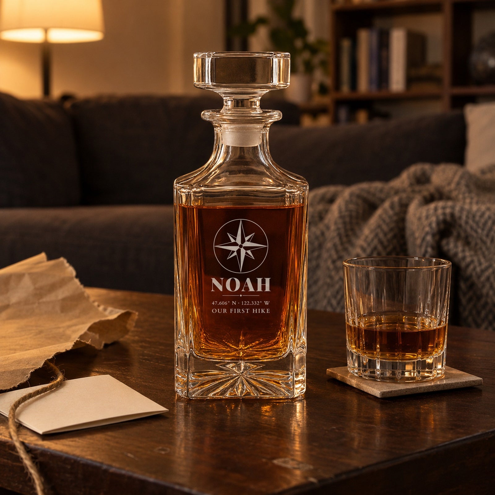 Personalized decanter for boyfriend on a coffee table beside torn kraft wrapping paper, engraved Noah with Seattle coordinates and Our First Hike