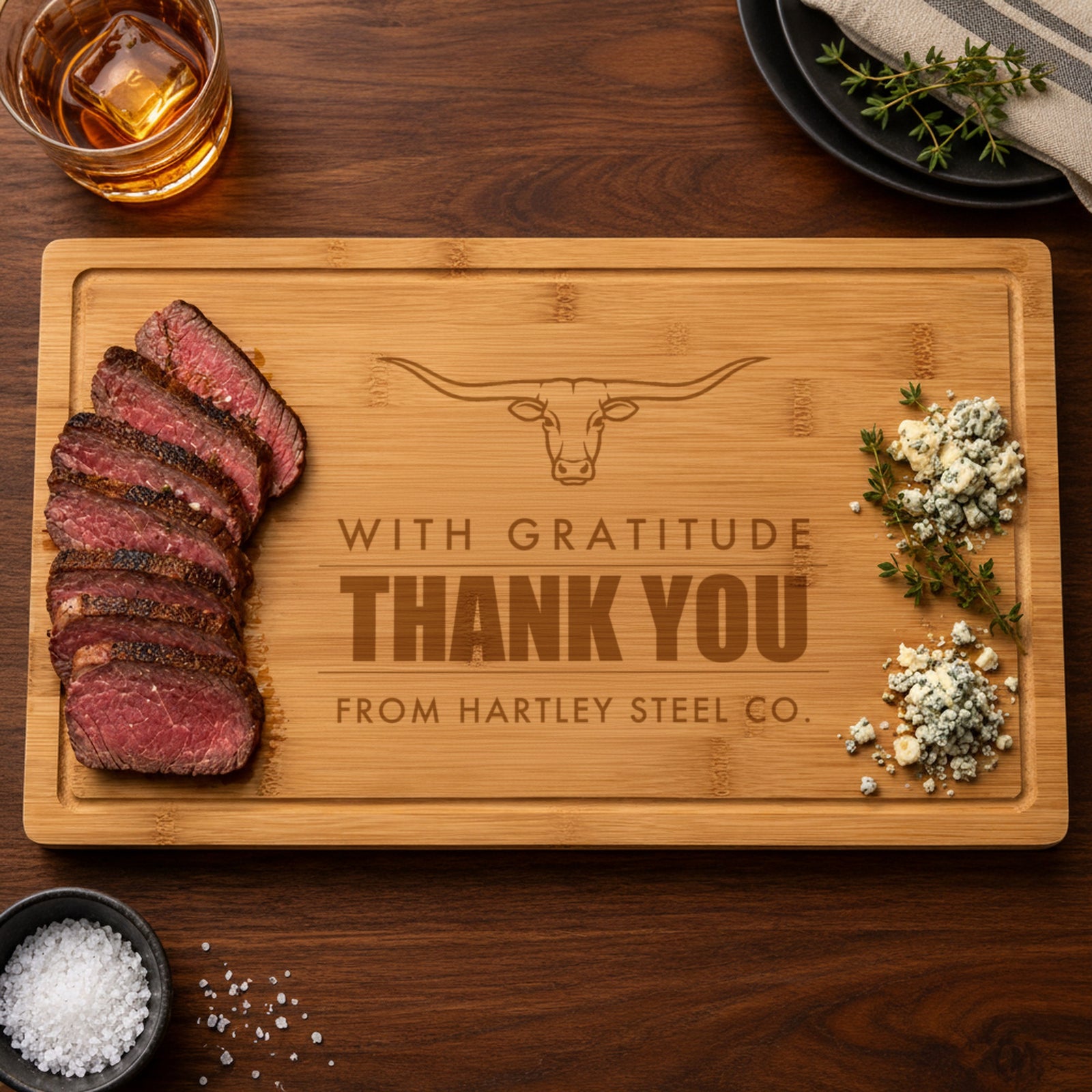 Engraved client gift cutting board reading With Gratitude, Thank You and From Hartley Steel Co., served with sliced steak, blue cheese and herbs