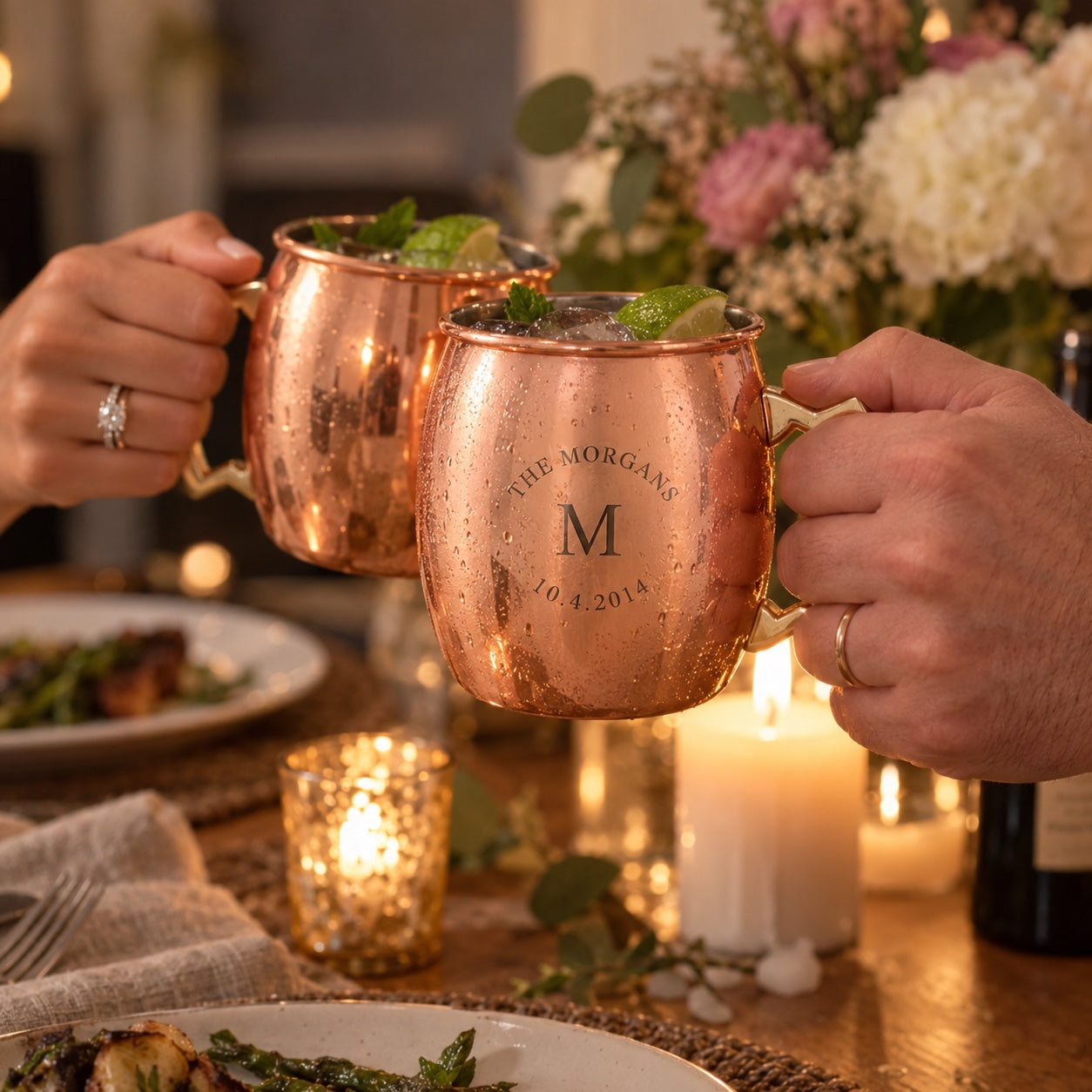 A husband and wife clink personalized copper mugs engraved THE MORGANS, M and 10.4.2014 over a candlelit anniversary dinner table at home.