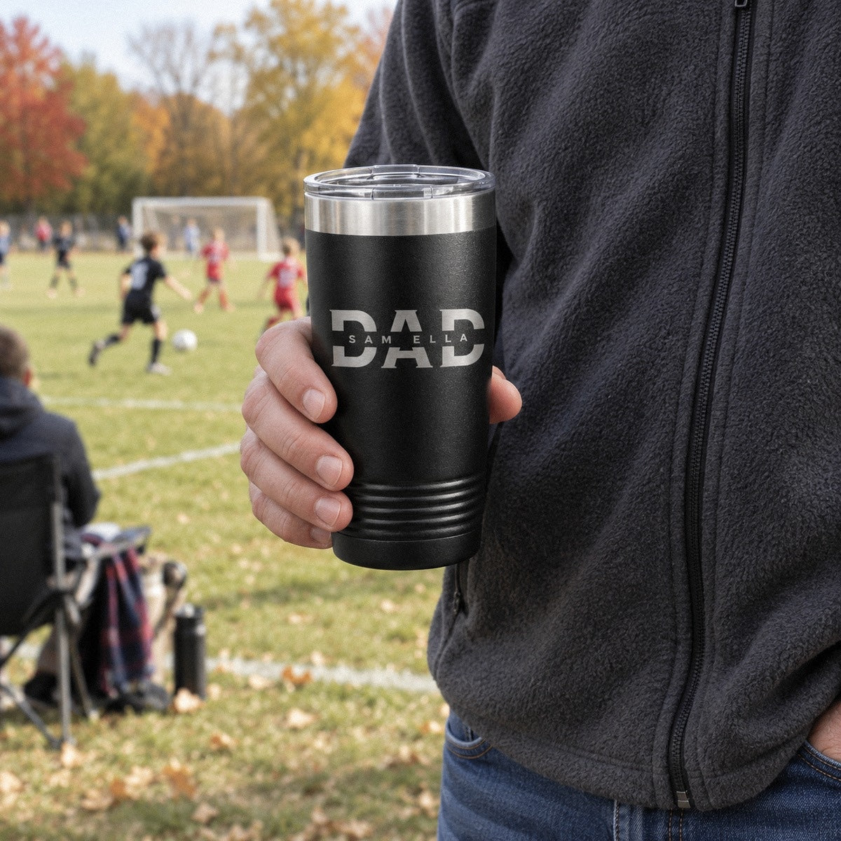 A dad holds his personalized dad tumbler of coffee at his kids' soccer game, engraved with a big DAD and the names SAM and ELLA