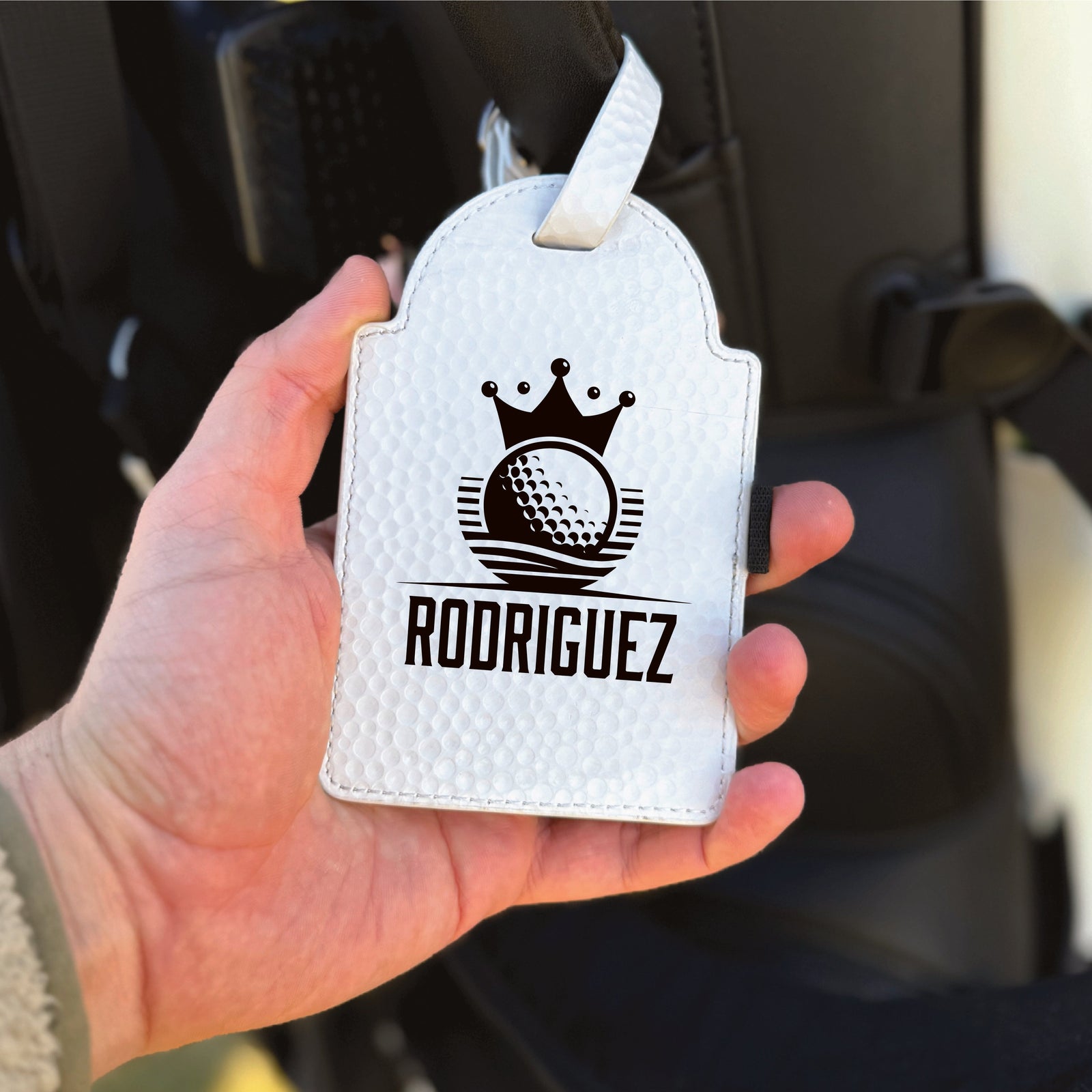 White leather golf towel tag with crown and golf ball motif, personalized with sample name "RODRIGUEZ" in black engraving, shown held in hand with white stitching detail and attachment loop