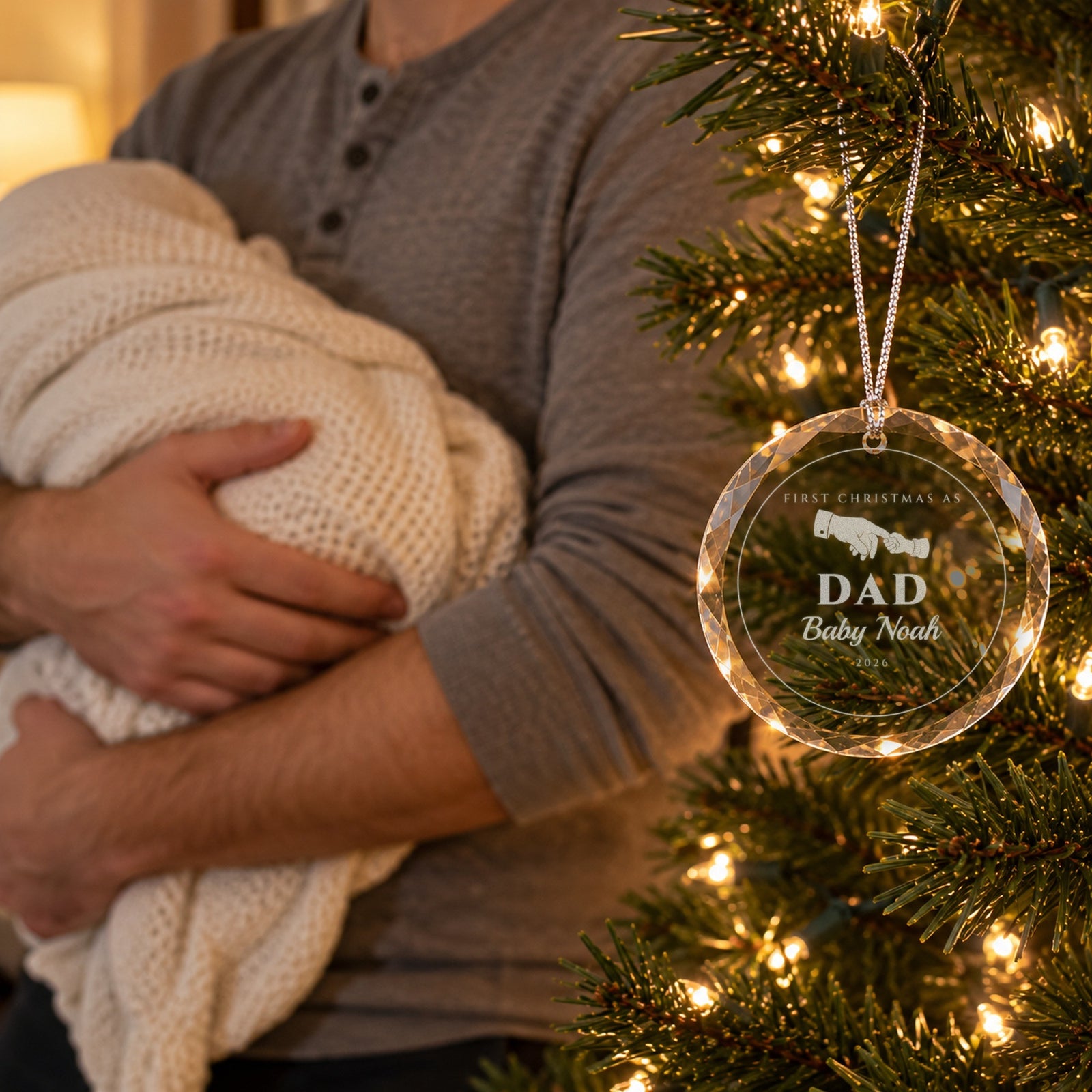 New dad holding his sleeping newborn by the Christmas tree beside the engraved ornament reading FIRST CHRISTMAS AS DAD, Baby Noah, 2026