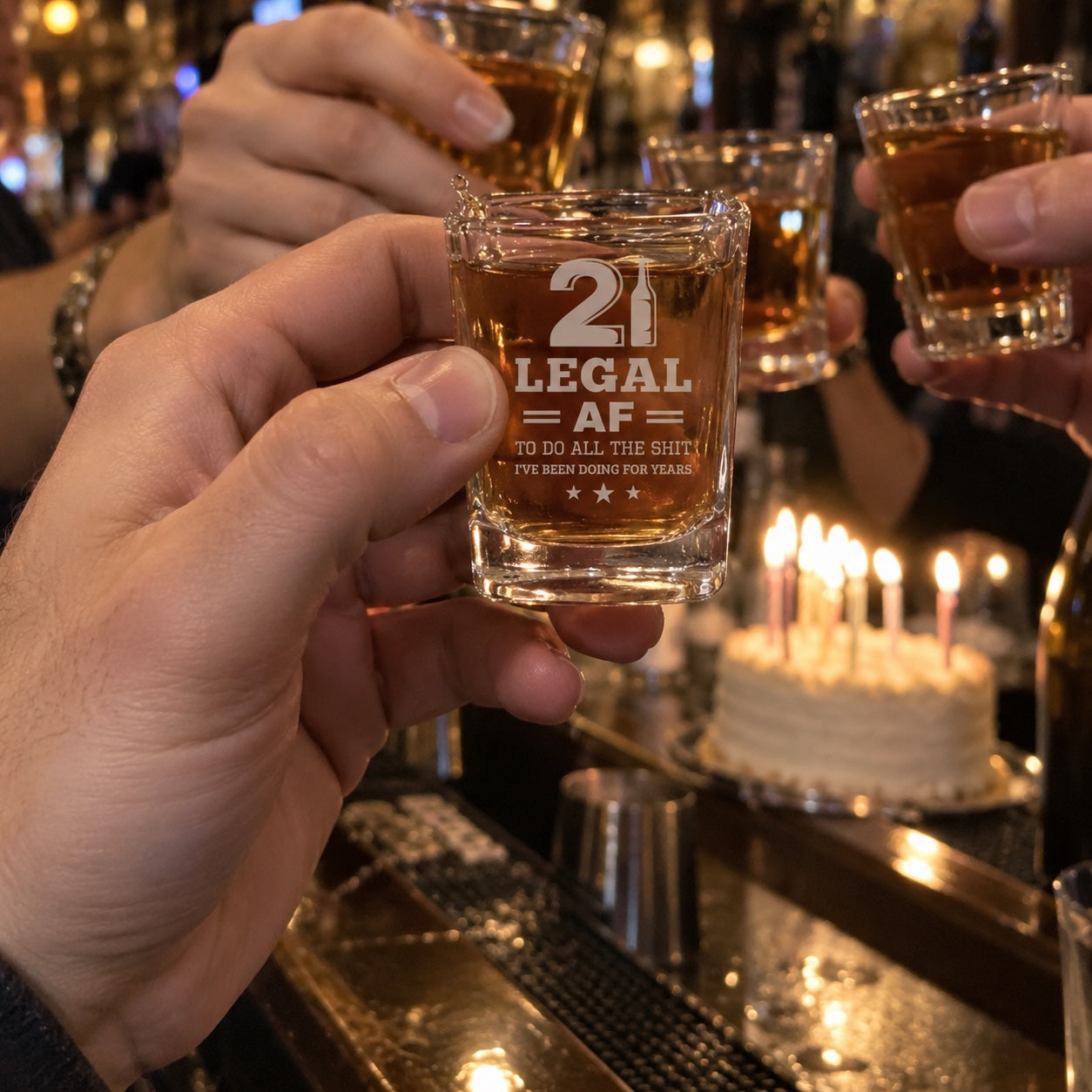 Friends raising shots at a 21st birthday at the bar, the front Legal AF birthday shot glass sharp, a birthday cake with lit candles behind