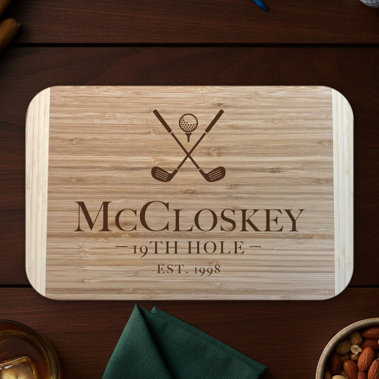 Personalized 19th Hole cutting board on a dark walnut bar top with golf balls, tees and whiskey, engraved McCloskey 19TH HOLE EST. 1998 under crossed clubs on two tone bamboo.