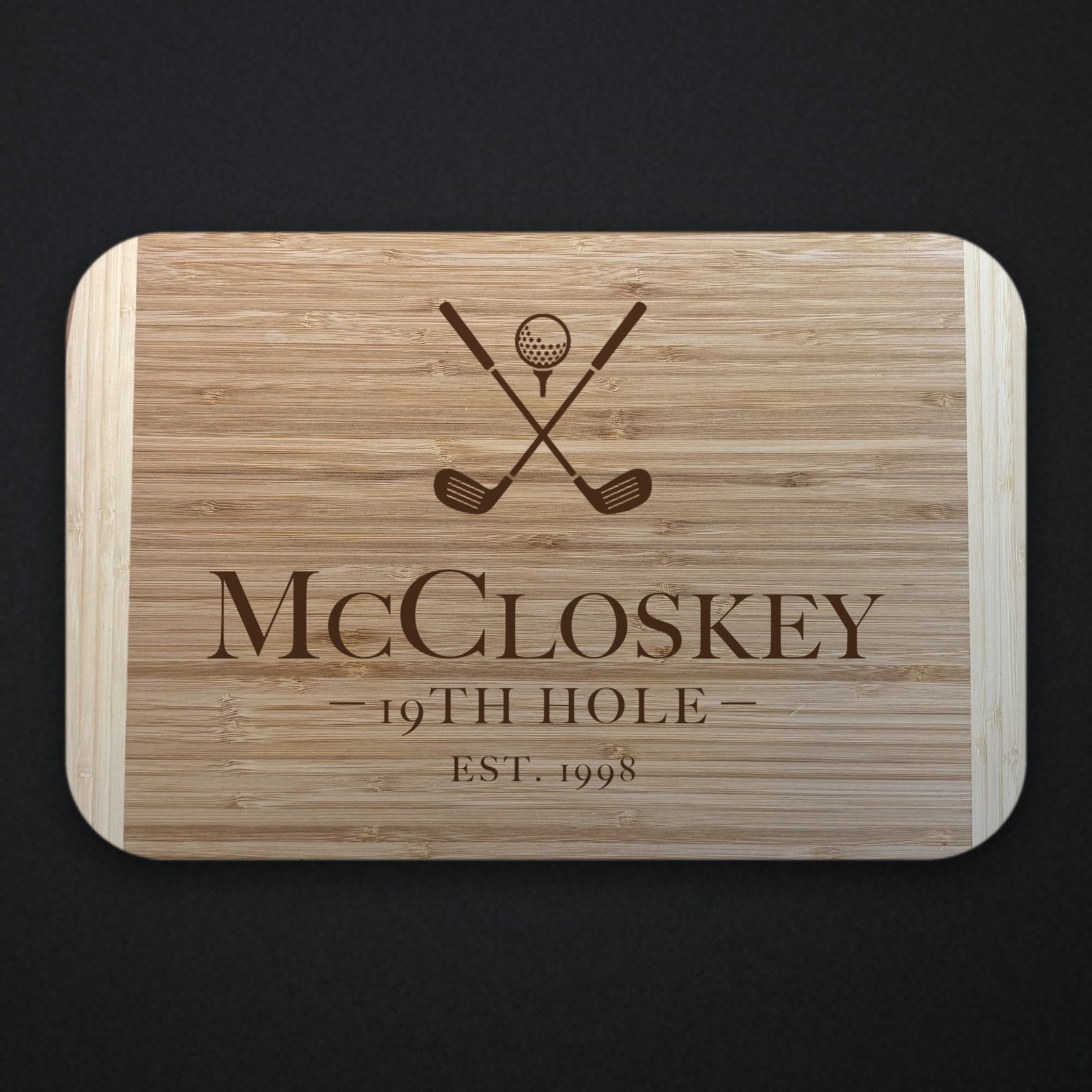 Two tone bamboo cutting board engraved with crossed golf clubs and a ball above the name McCloskey, the line 19TH HOLE and EST. 1998, shown on a dark slate background.