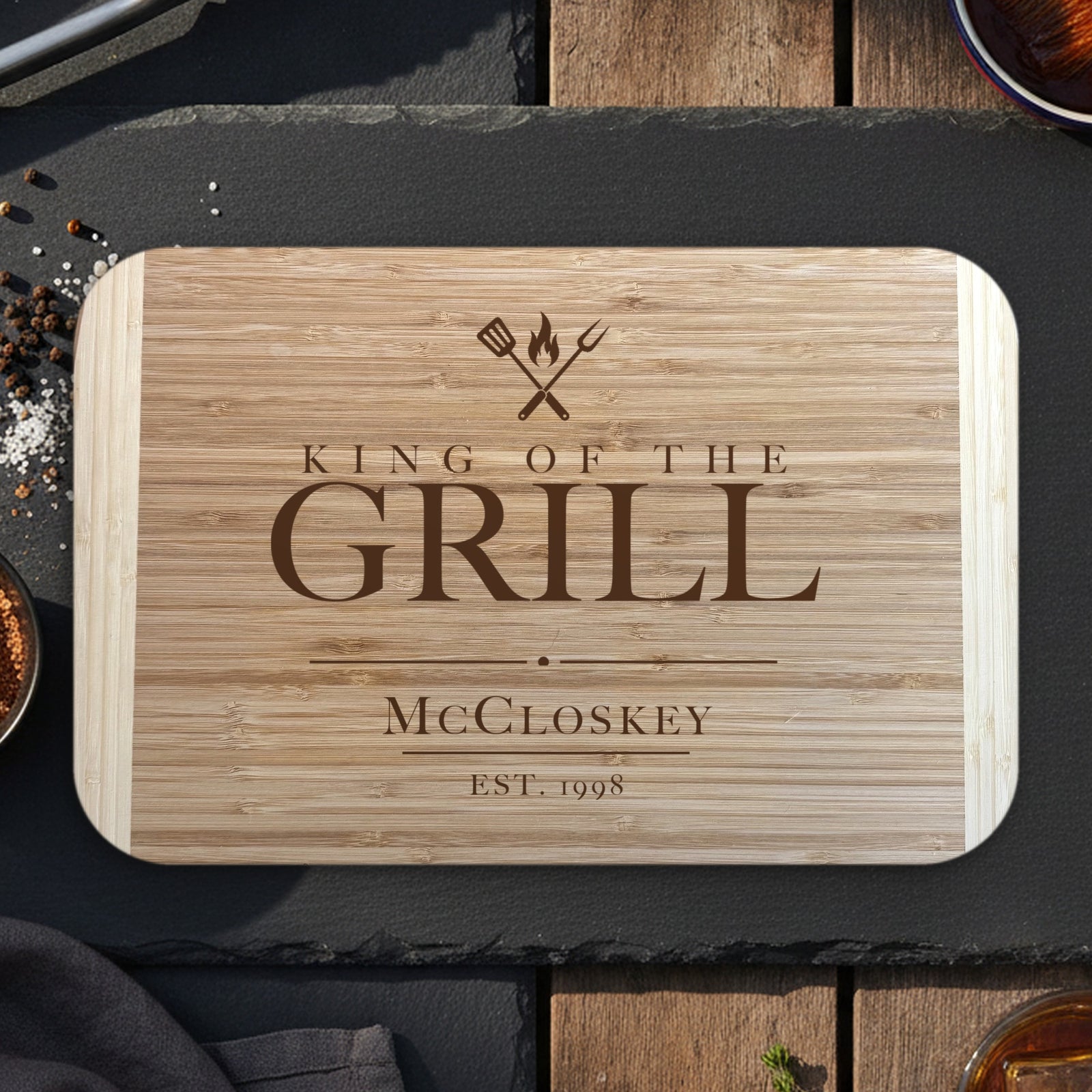 Personalized King of the Grill cutting board on dark slate with grilling tongs, dry rub and bourbon, engraved McCloskey EST. 1998 under crossed grill tools on two tone bamboo.