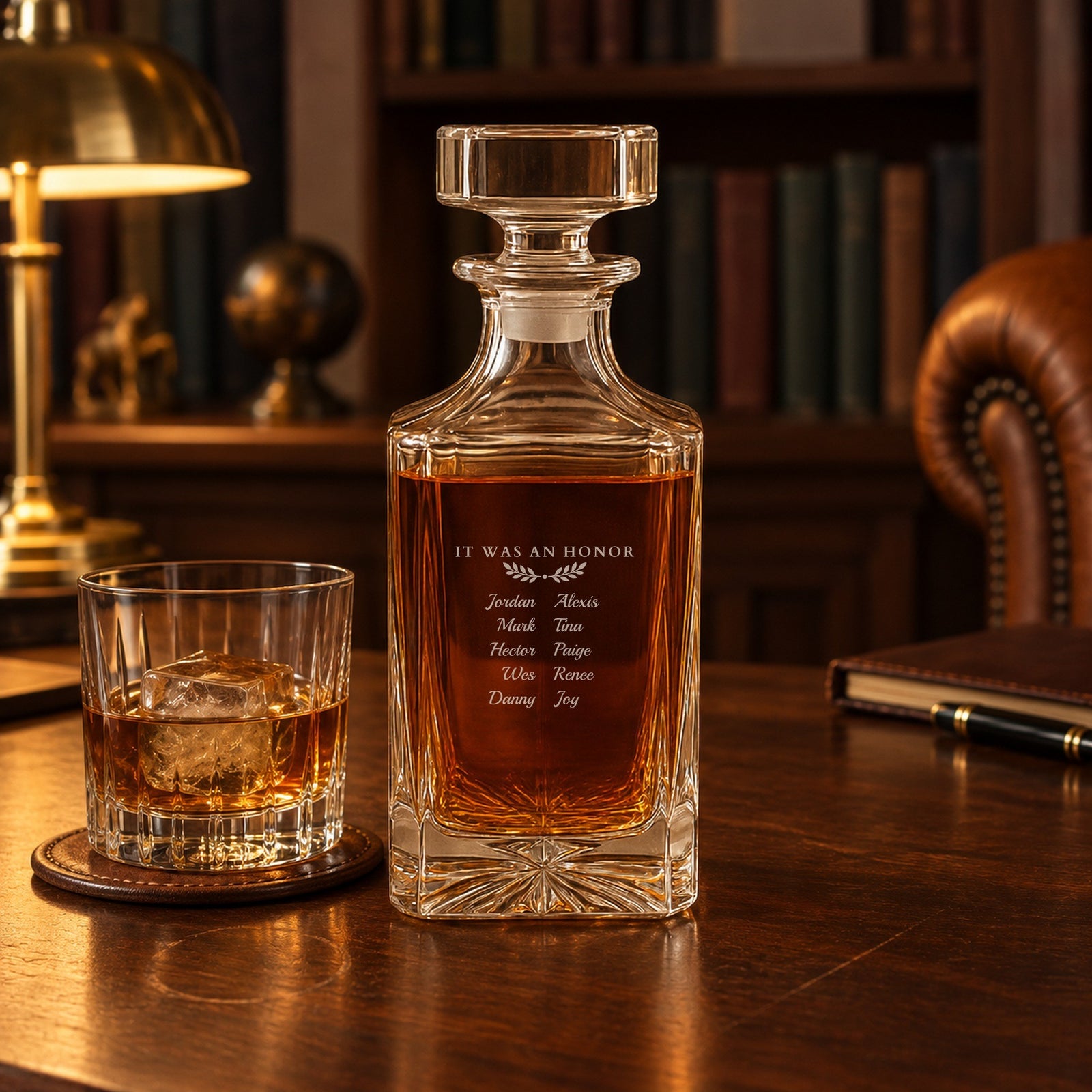 Gift for a departing boss in a home study: a whiskey decanter engraved It Was an Honor with ten team names, beside a rocks glass of bourbon