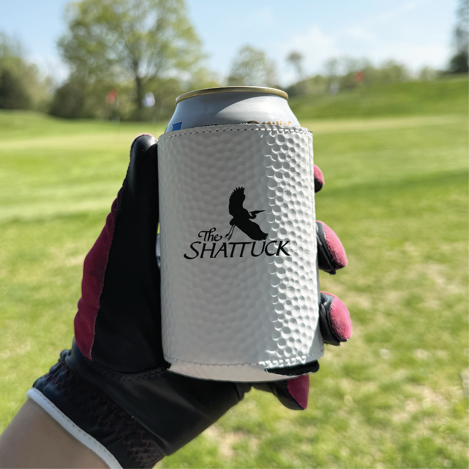 White golf ball-textured can cooler sleeve with "The SHATTUCK" engraved design and eagle motif, held in gloved hand on golf course background