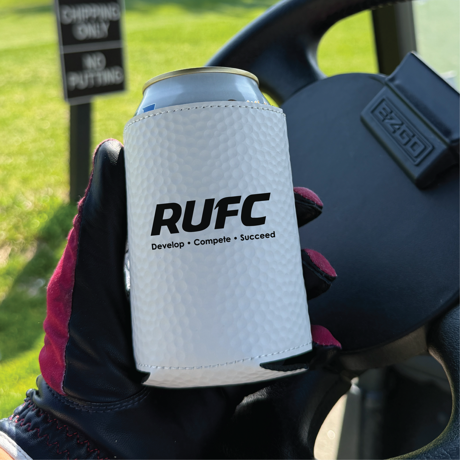 White golf ball textured can cooler sleeve with "RUFC Develop • Compete • Succeed" text, held in gloved hand on golf course with cart in background