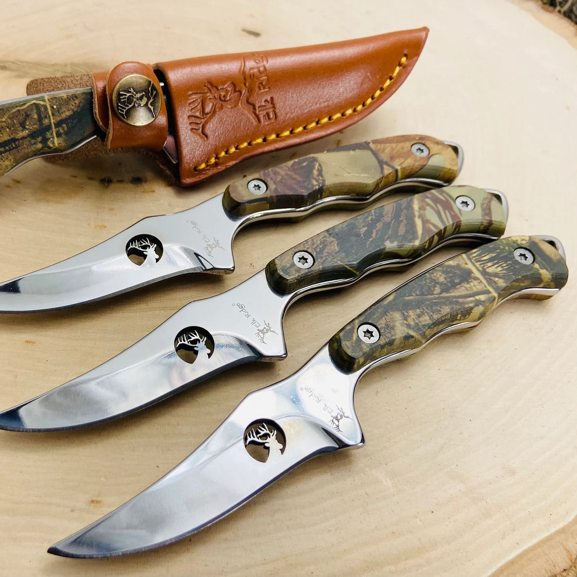 Three fixed-blade knives with camouflage wood handles and engraved stainless steel blades featuring elk head motif, displayed with matching brown leather sheath with decorative stitching
