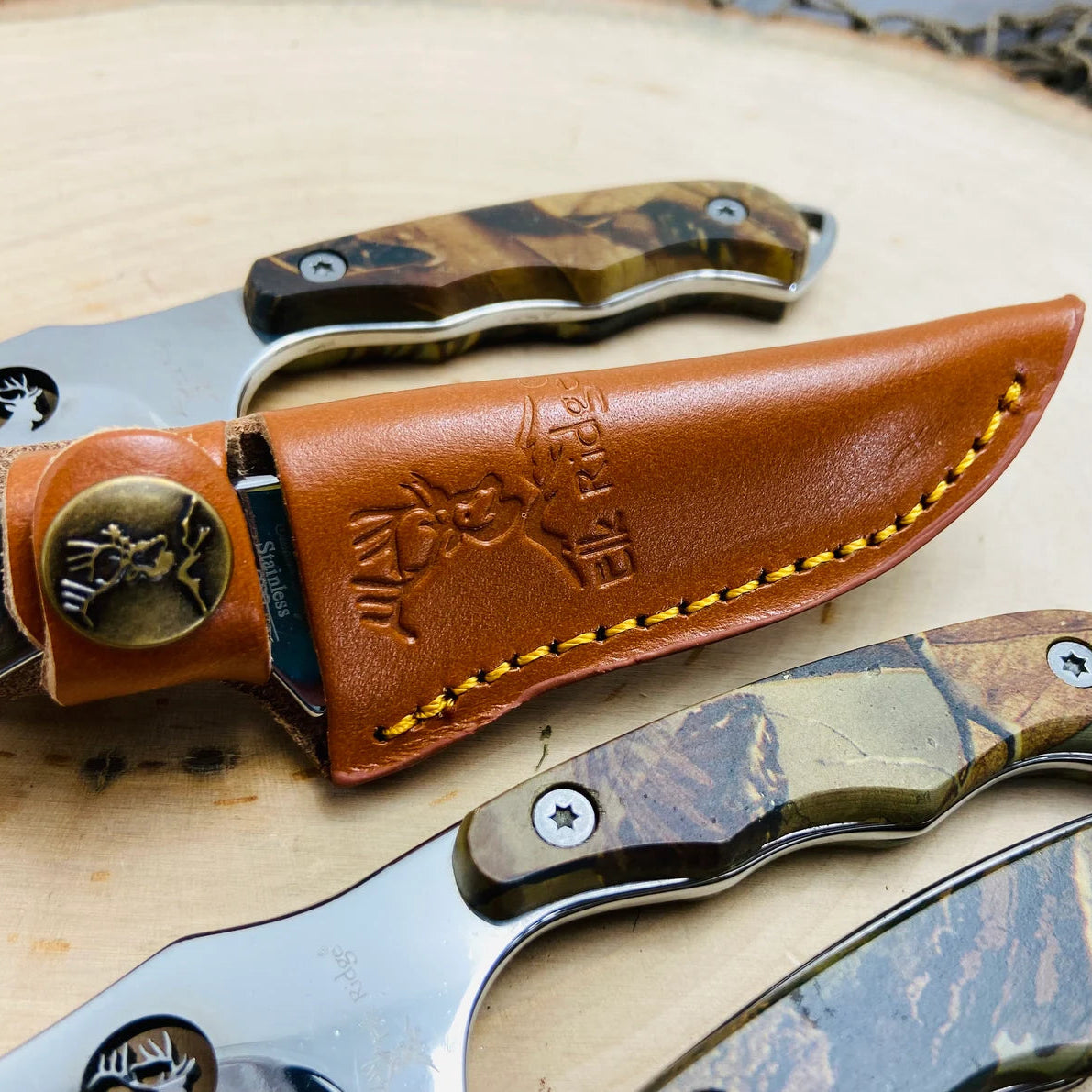 Tan leather sheath with brass hardware and yellow stitching, displaying embossed design and metal medallion, paired with three fixed-blade knives with wood handles