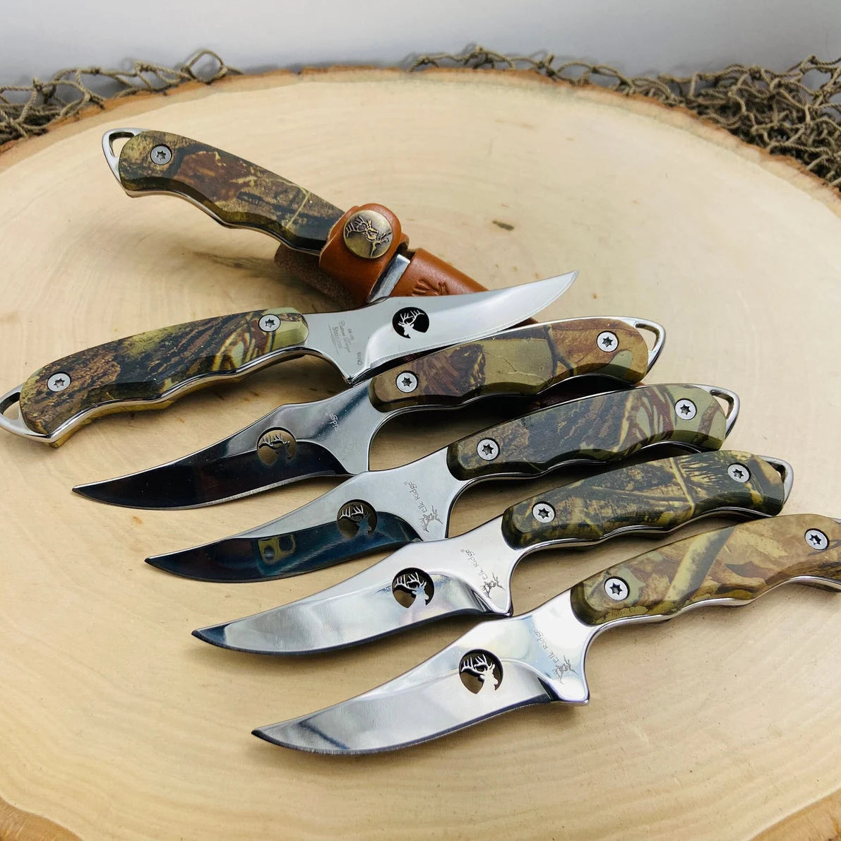 Collection of six fixed blade hunting knives with camouflage handles and stainless steel blades, each featuring a deer head emblem, displayed on a natural wood surface with decorative twine.