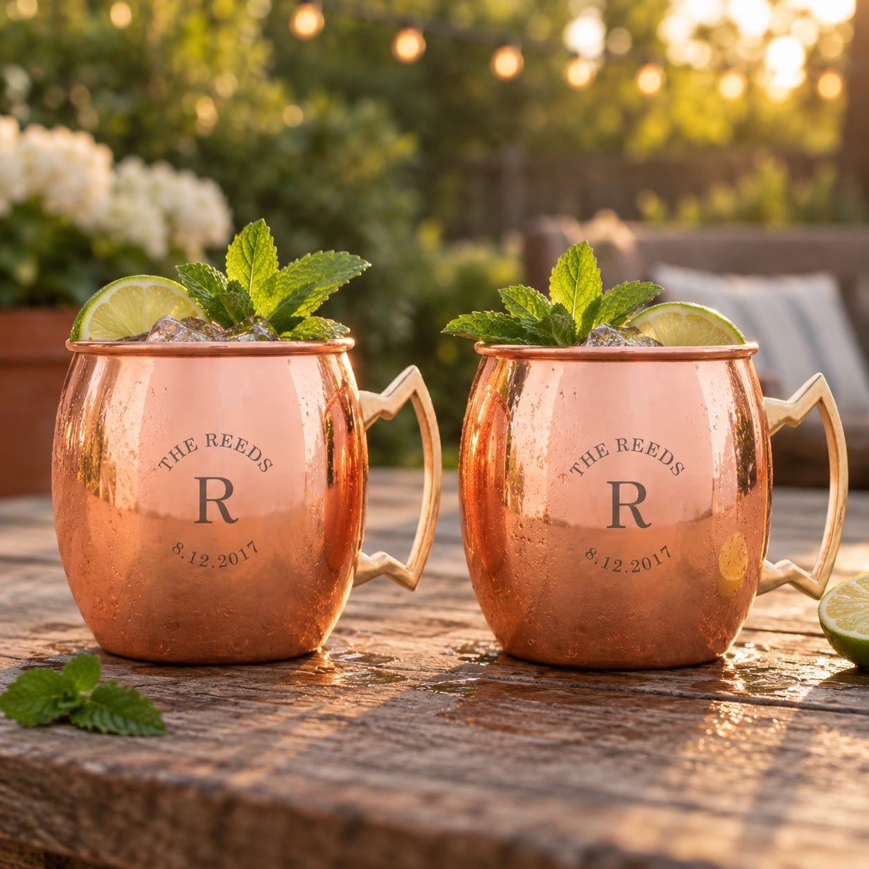 A matching pair of couple's copper Moscow mule mugs both engraved THE REEDS, R and 8.12.2017, filled with mules on a patio table at golden hour.