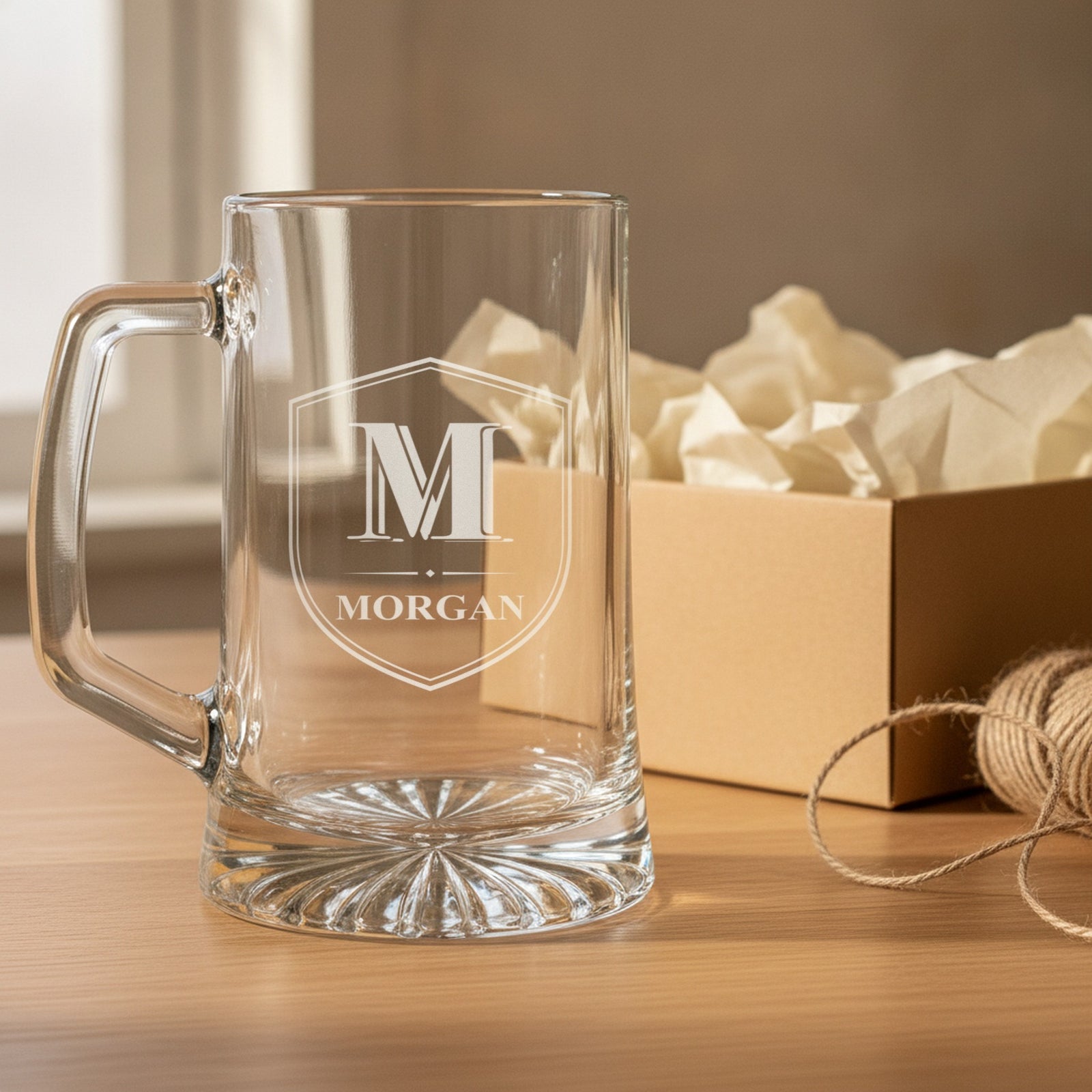 Empty personalized monogram beer stein beside an open kraft gift box with tissue paper, etched with a Shield Crest reading M and the name Morgan