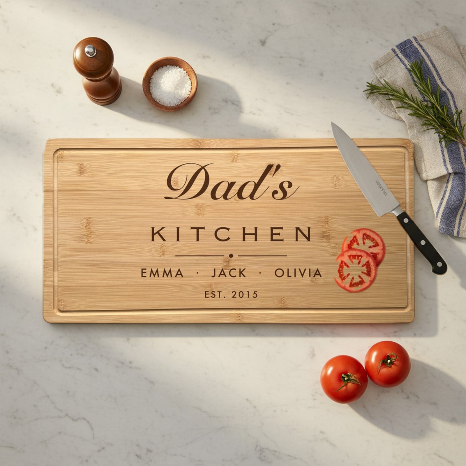 Engraved Dad's Kitchen bamboo board on a marble kitchen counter with a chef knife, sliced tomatoes, a pepper mill, sea salt and a striped dish towel.