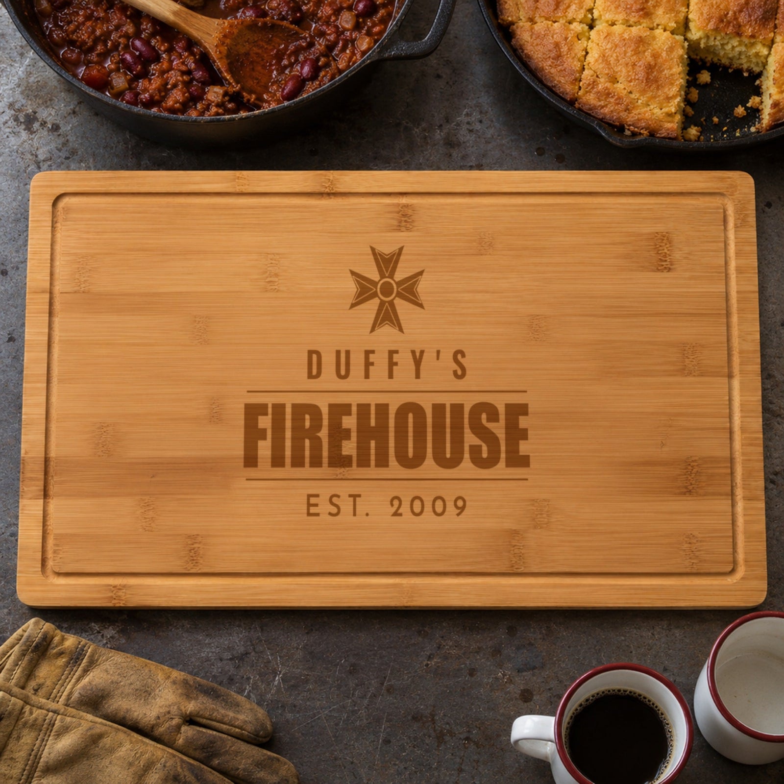 Personalized firehouse cutting board engraved with a Maltese cross, DUFFY'S FIREHOUSE EST. 2009, beside a pot of chili and cornbread, a gift for firefighters