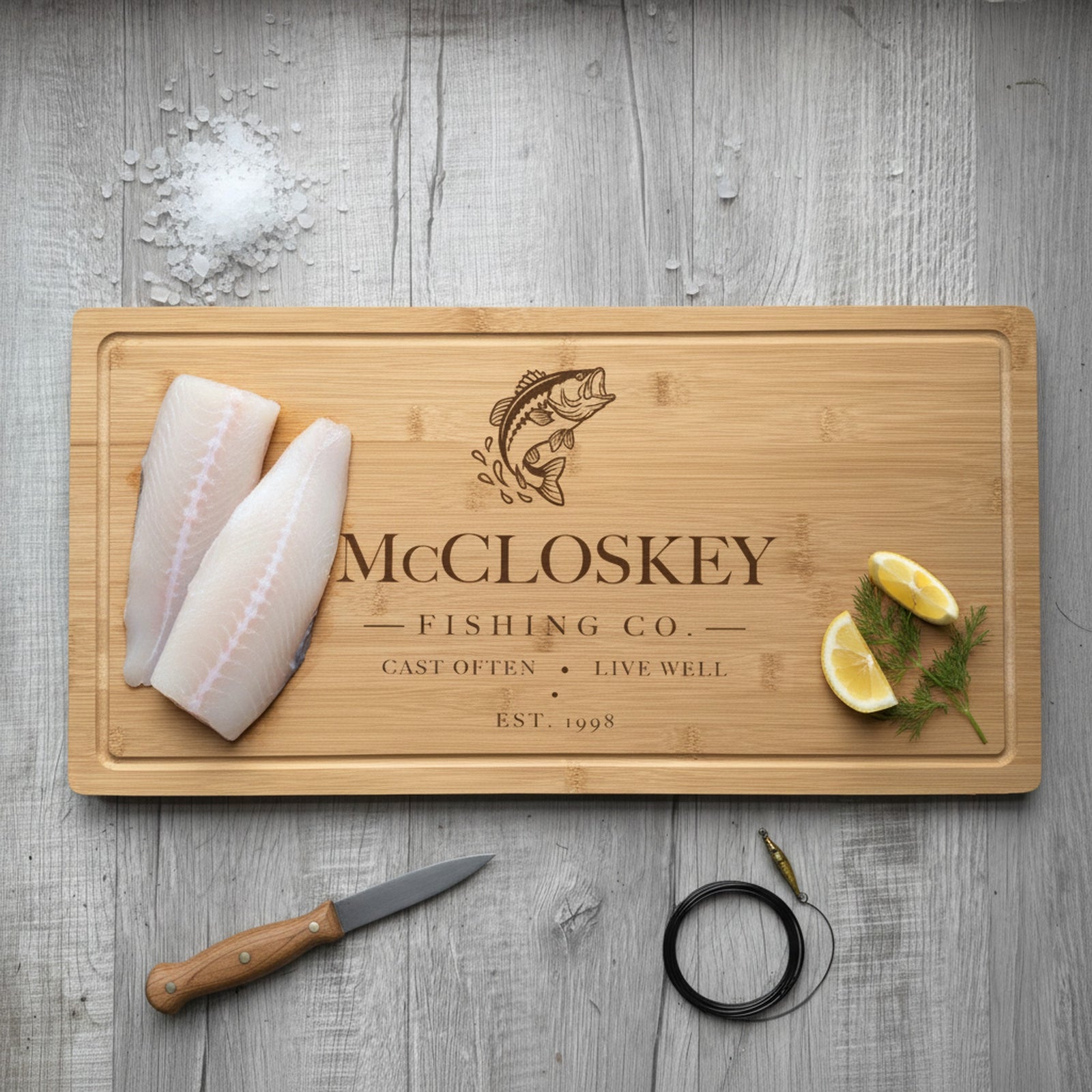 Engraved fishing bamboo board on weathered dock planking with two fresh fish fillets at one end, lemon wedges and dill at the other, beside a fillet knife.