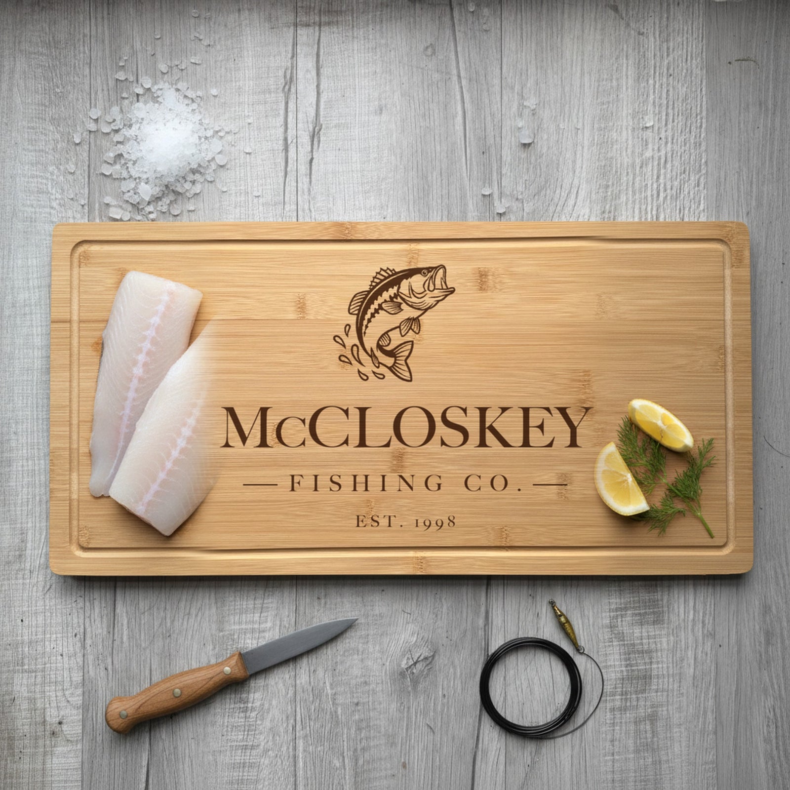 Engraved fishing bamboo board on weathered dock planking with two fresh fish fillets at one end, lemon wedges and dill at the other, beside a fillet knife.