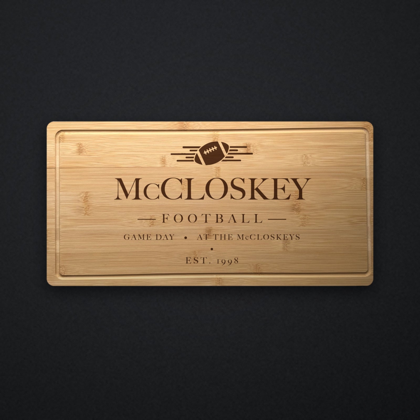 Personalized football tailgate cutting board in solid bamboo on dark slate, engraved with a football and speed lines above the family name and the year.
