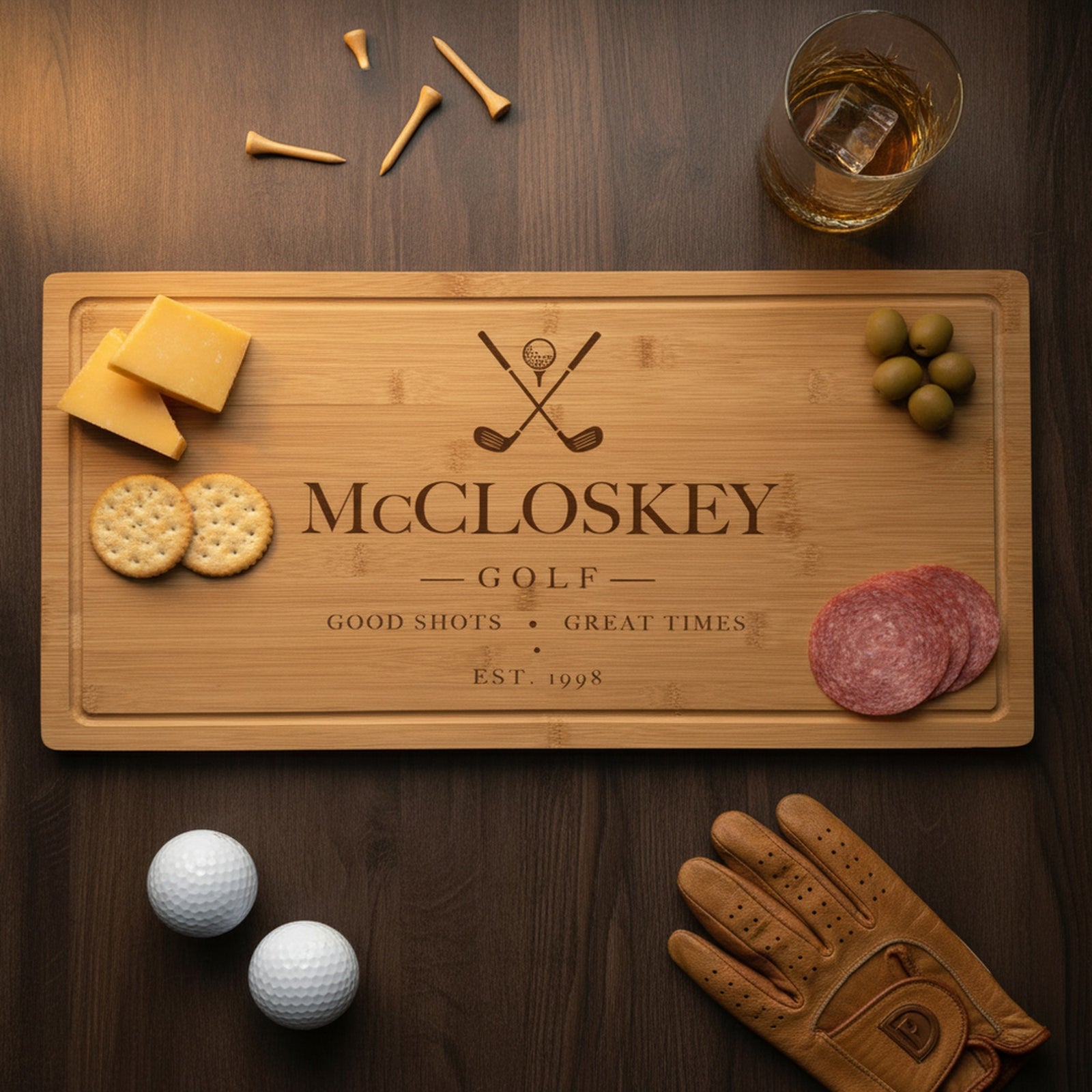 Engraved golf bamboo board on a dark table with cheddar and crackers at one end, olives and salami at the other, beside a whiskey glass, golf balls and a leather glove.