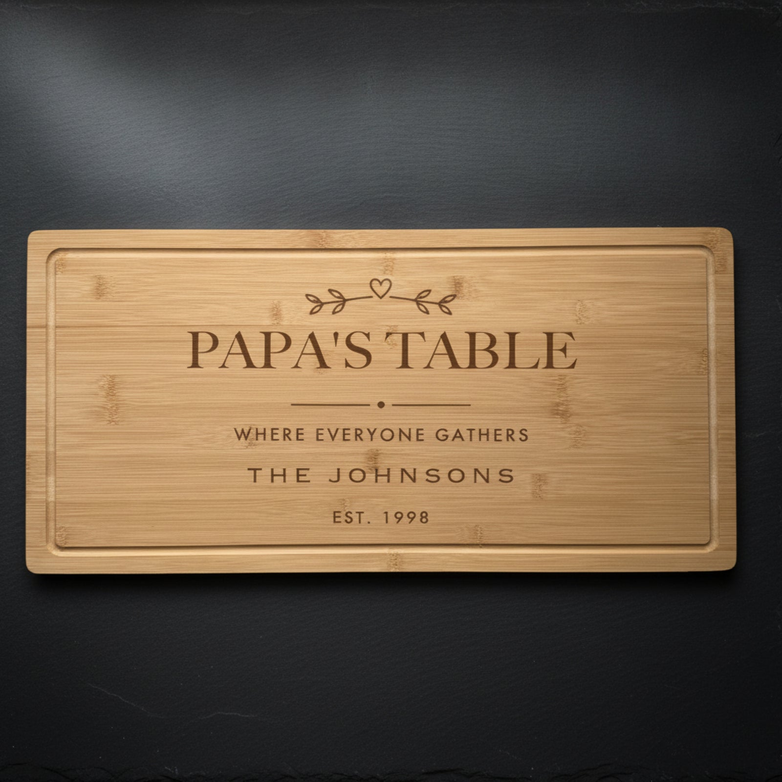 Personalized grandpa cutting board in solid bamboo on dark slate, engraved Papa's Table above the family name and the year, with a small sprig and heart.