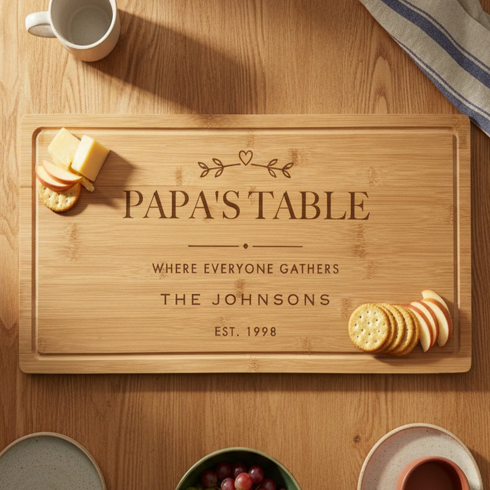 Engraved Papa's Table bamboo board on a warm oak family table with mugs, plates, grapes and cheese and crackers set at each end of the board.