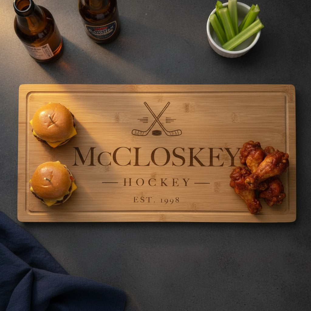Engraved hockey bamboo board on a slate counter with sliders at one end and chicken wings at the other, beside beer bottles and celery on game night.
