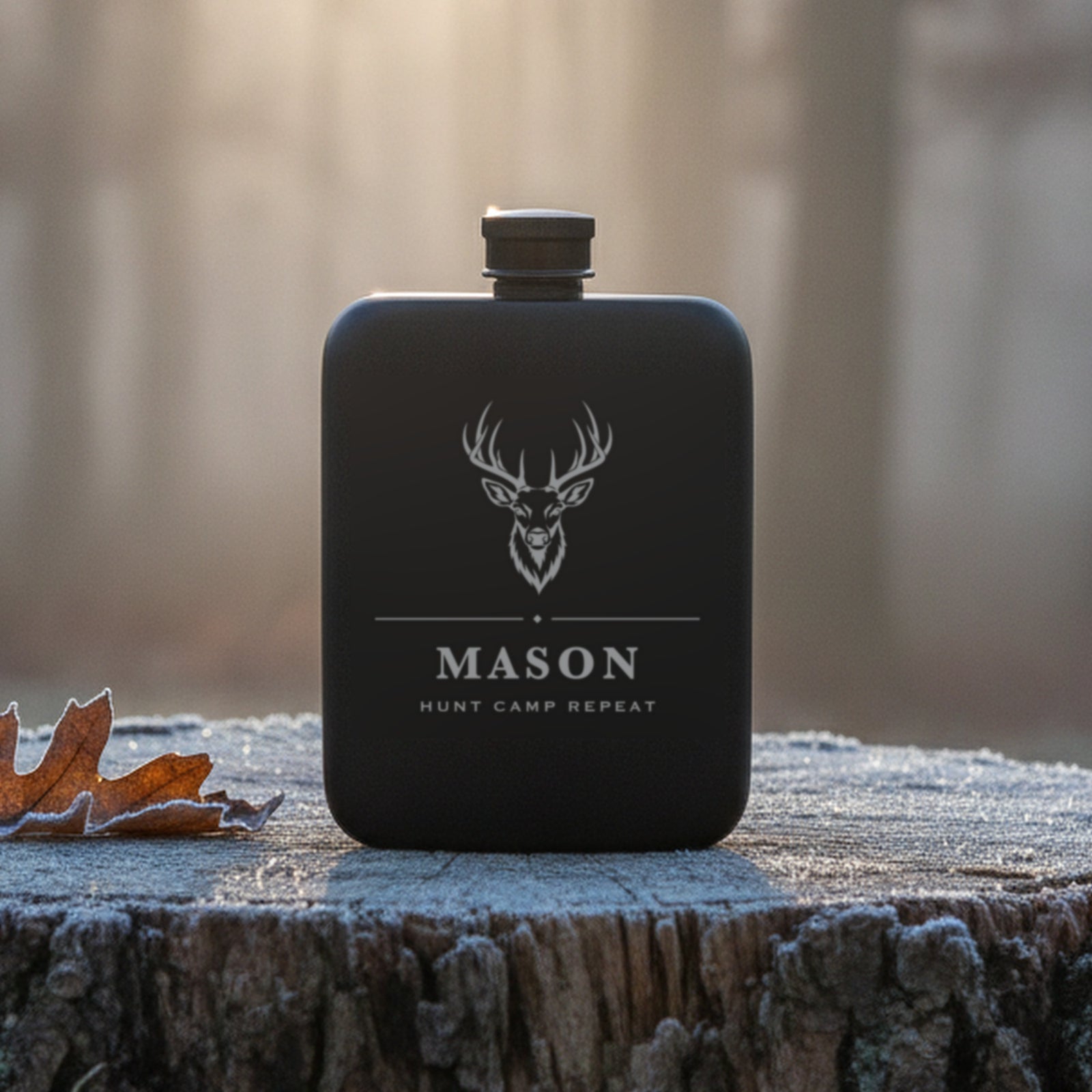 Personalized buck engraved hunting flask reading Hunt Camp Repeat, standing on a frosted oak stump at first light in misty woods.