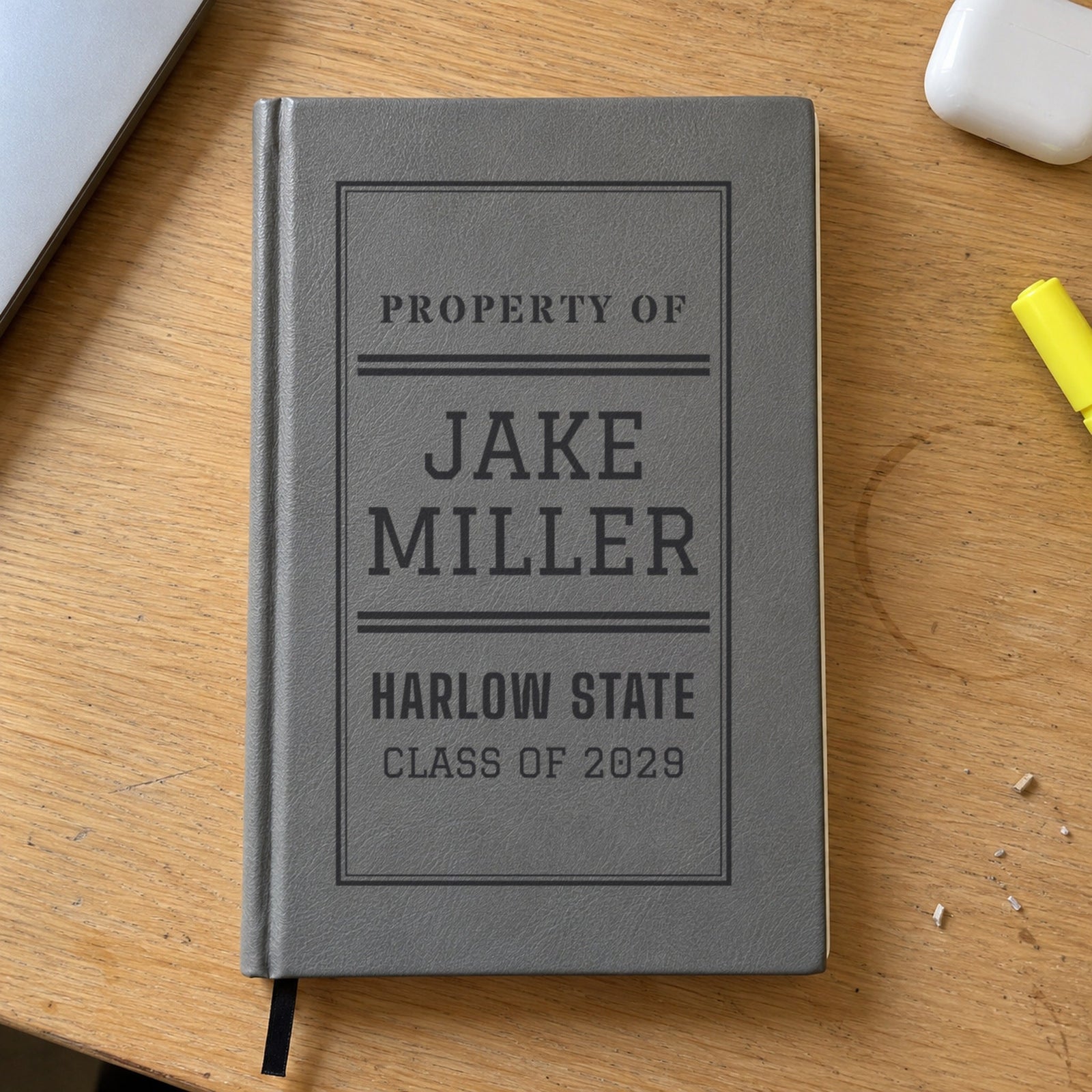 Personalized journal for a college student in gray engraved Property Of Jake Miller, Harlow State, Class of 2029, on a dorm desk with a laptop and highlighter