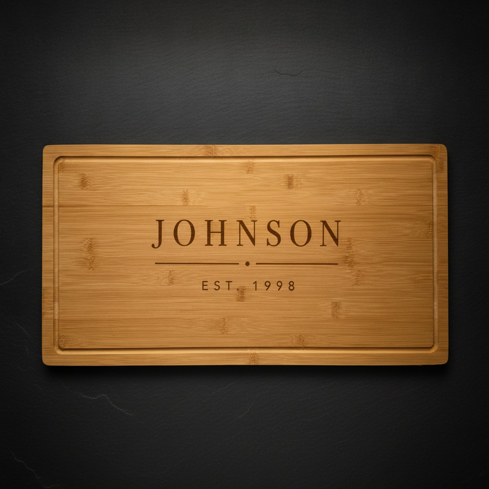 Personalized last name cutting board in solid bamboo on dark slate, laser engraved JOHNSON above EST 1998 in clean block type.