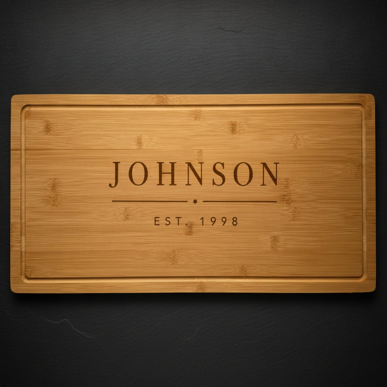 Personalized last name cutting board in solid bamboo on dark slate, laser engraved JOHNSON above EST 1998 in clean block type.