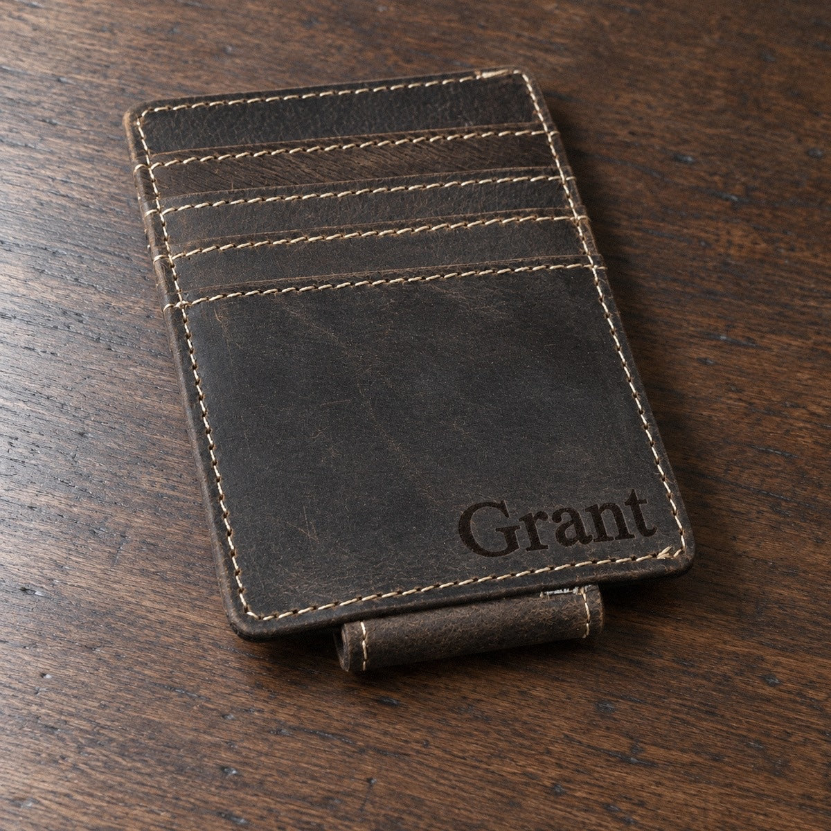 Personalized leather money clip wallet in dark distressed leather on a walnut desk, the name Grant engraved in the lower corner of the front