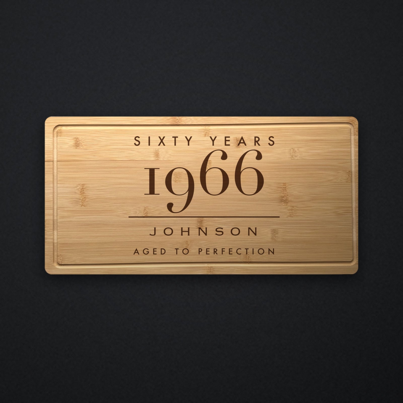 Personalized milestone birthday cutting board in solid bamboo on dark slate, engraved Sixty Years above the birth year, the name and Aged To Perfection.