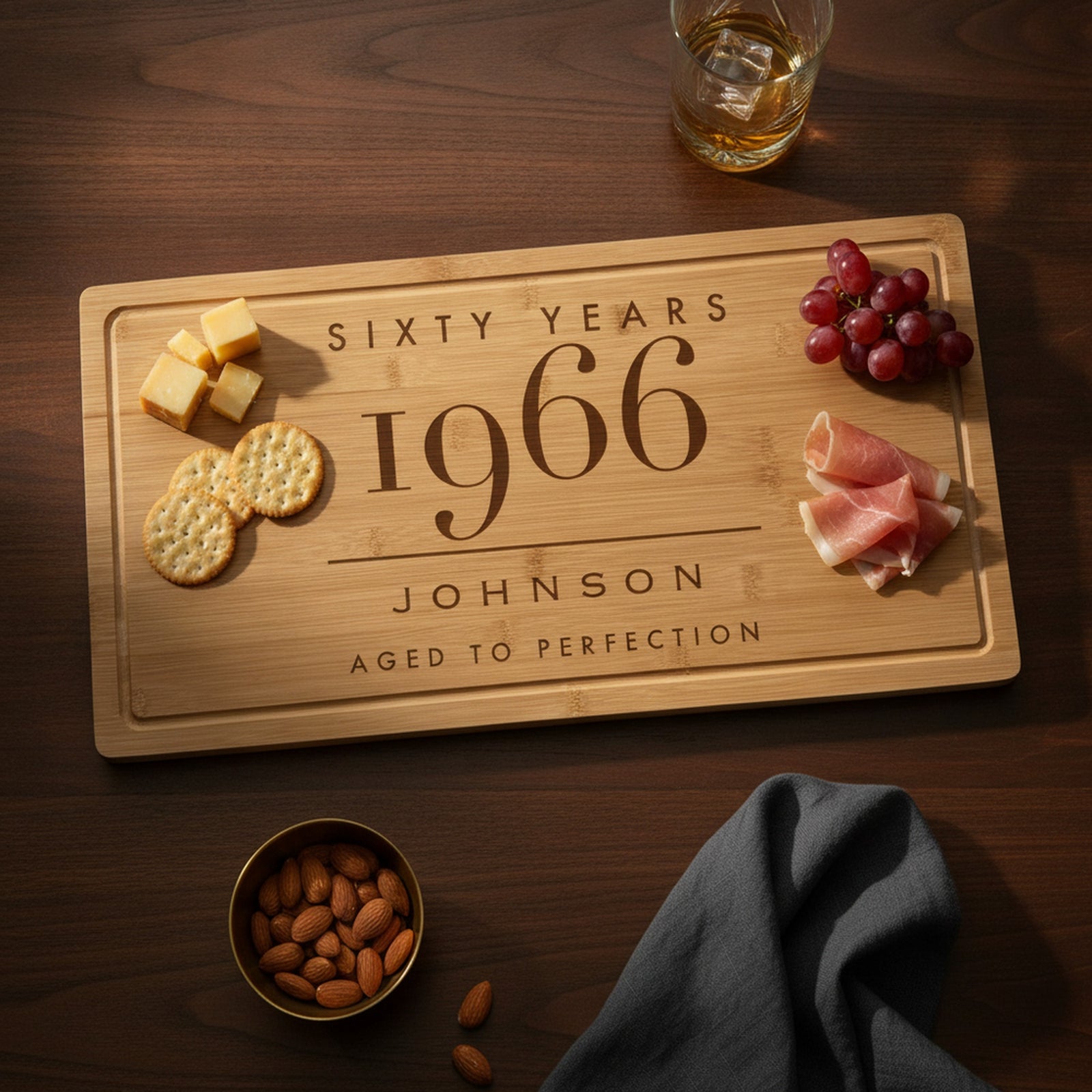 Engraved milestone birthday bamboo board on a dark walnut table with aged cheese and crackers at one end, grapes and prosciutto at the other, beside a whiskey glass.