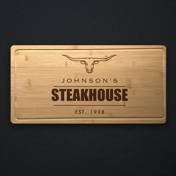 Personalized steakhouse cutting board in solid bamboo on dark slate, engraved with a longhorn steer above the name and the word Steakhouse.