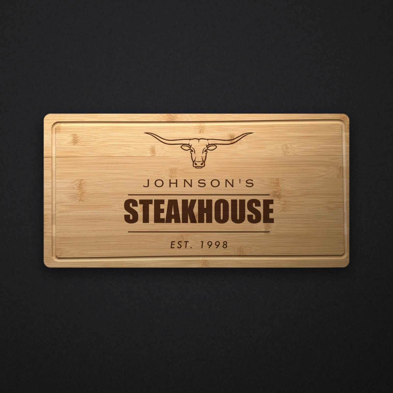 Personalized steakhouse cutting board in solid bamboo on dark slate, engraved with a longhorn steer above the name and the word Steakhouse.
