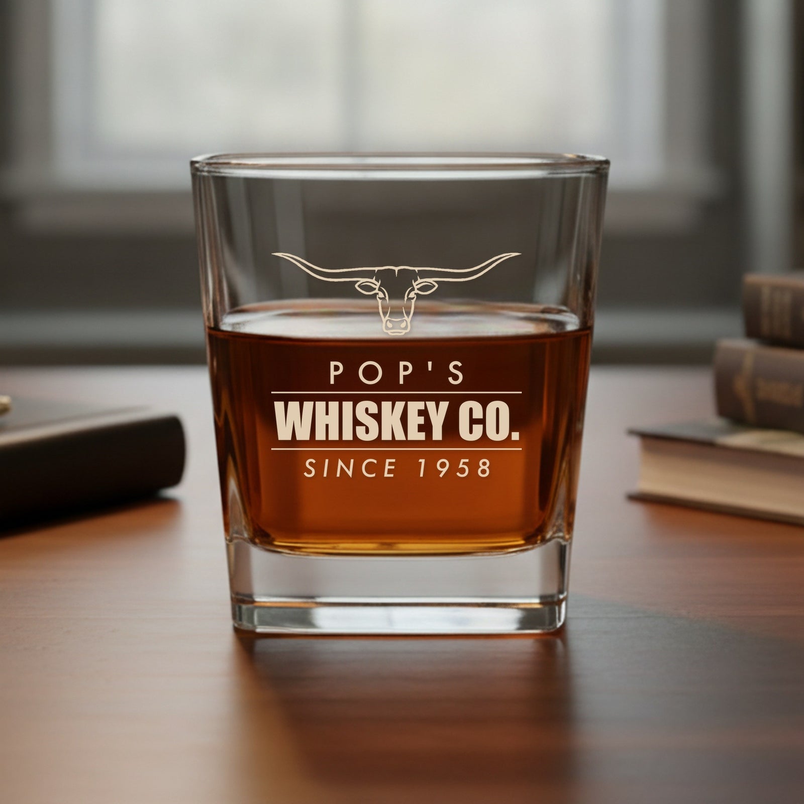 Engraved whiskey glass reading Pop's Whiskey Co., Since 1958 under a longhorn, set on a study desk by a window with books nearby
