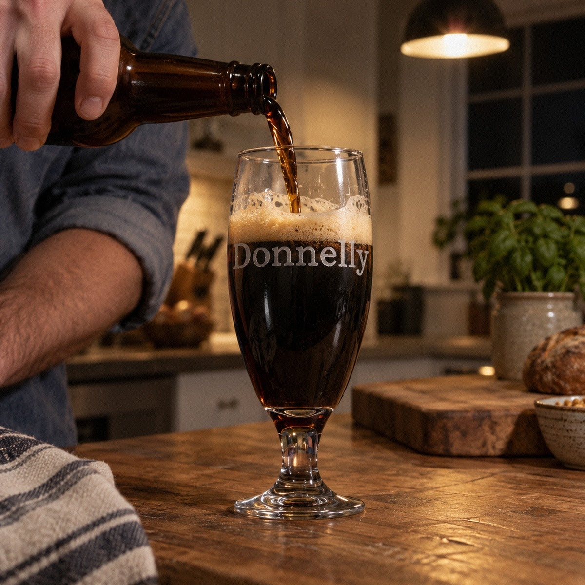 A man pours a dark stout into his personalized goblet engraved with the name Donnelly on a wooden kitchen island at night