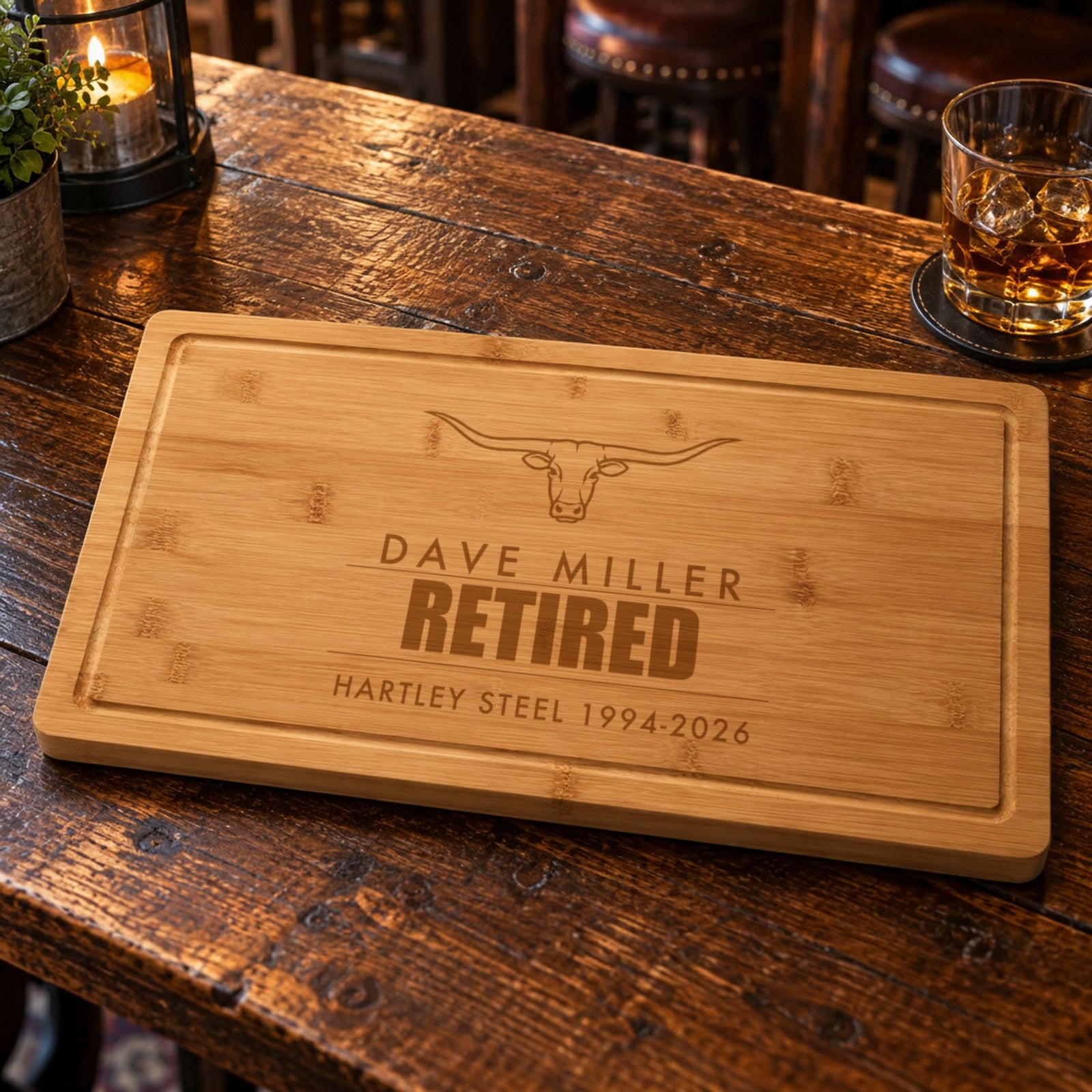 Bamboo retirement cutting board on a tavern table, engraved with Dave Miller above the word Retired and Hartley Steel 1994-2026 on the bottom line