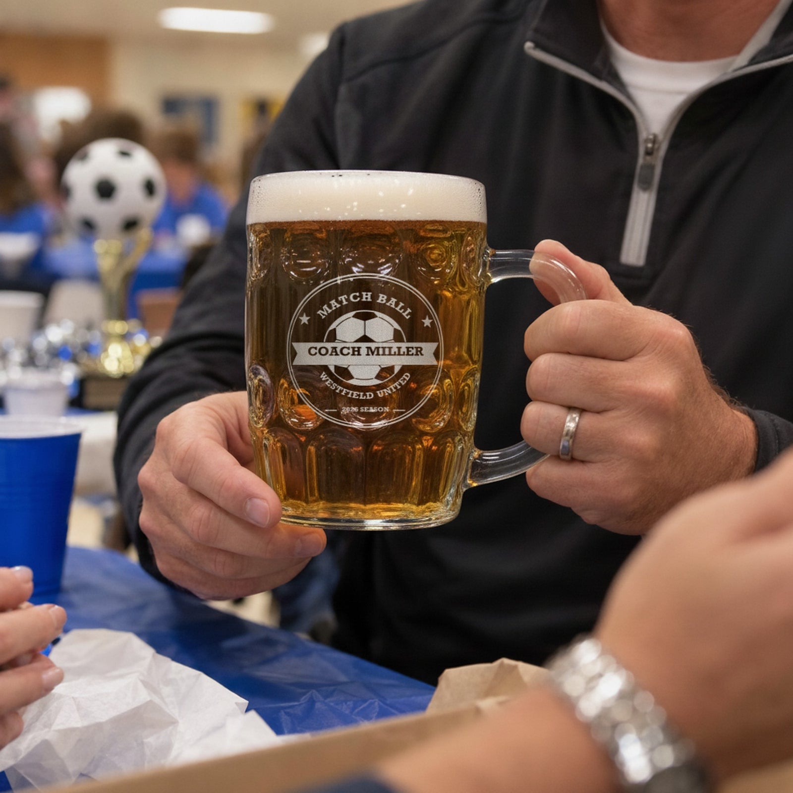 Soccer coach holding the personalized Match Ball beer stein at an end-of-season team banquet, with a soccer trophy in the background
