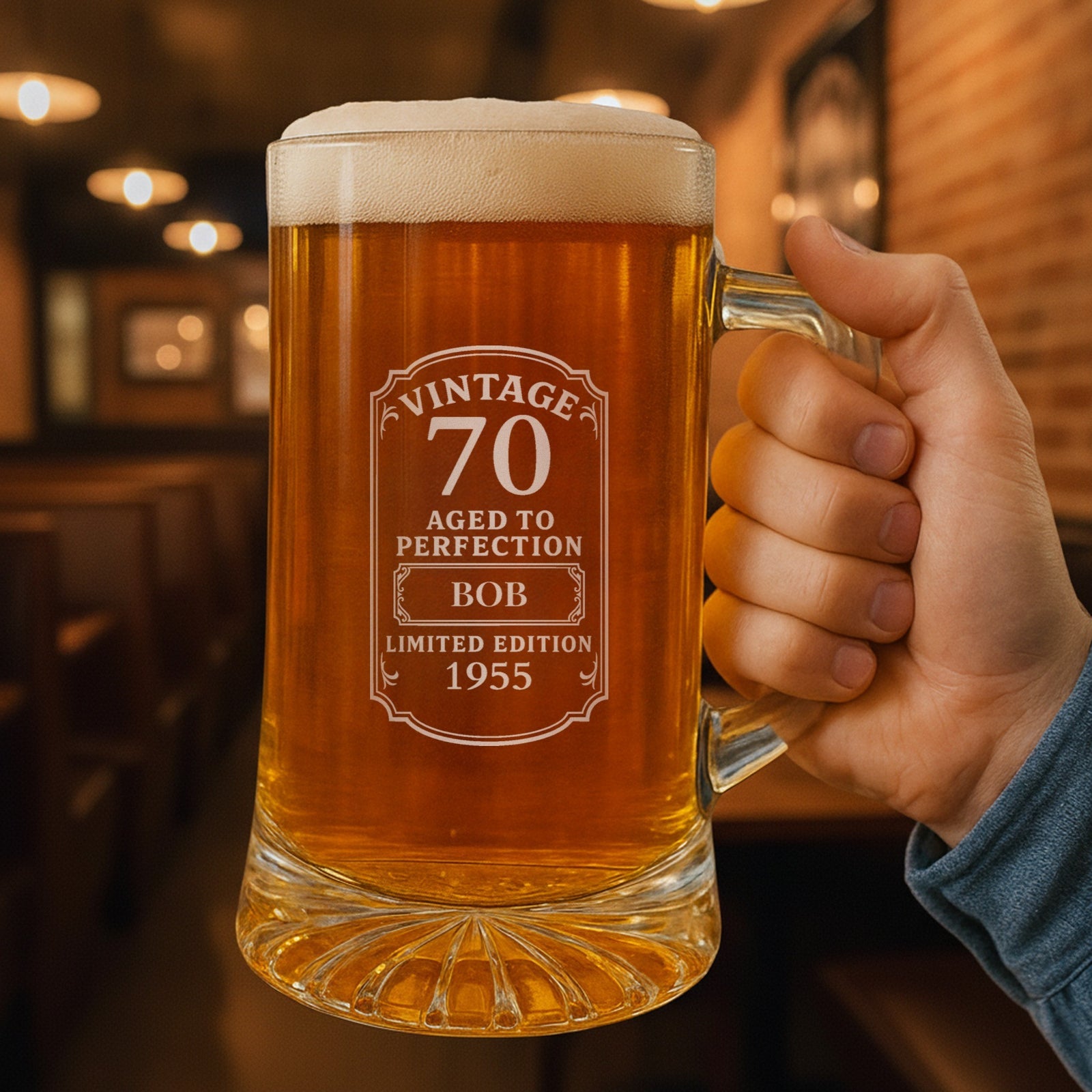 Hand holding a personalized 70th birthday beer stein in a warm pub, the Vintage 70 label engraved with the name Bob and the year 1955