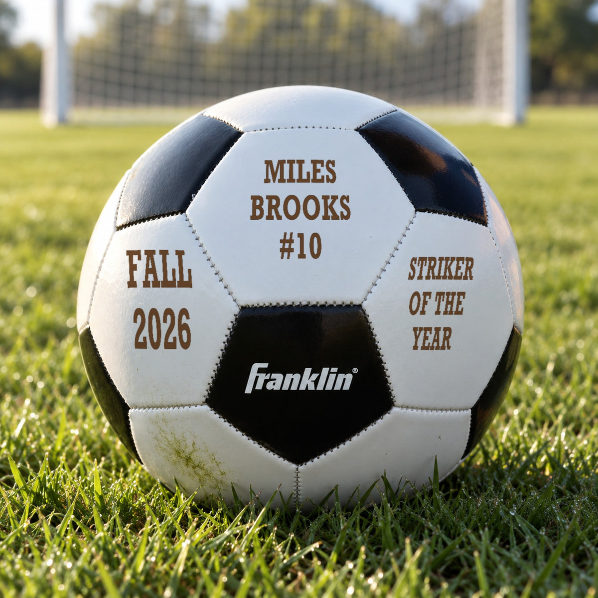 Personalized soccer ball engraved with a player's name and #10, Fall 2026 and Striker of the Year, resting in the grass in front of a goal