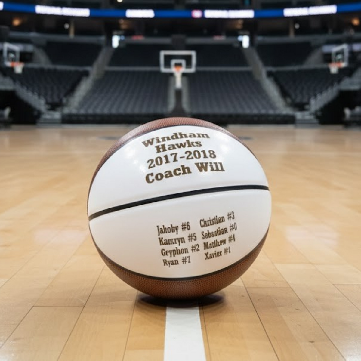 Basketball engraved with sample team name "Windham Hawks 2017-2018 Coach Win" and roster including player names and numbers on wooden court with arena in background