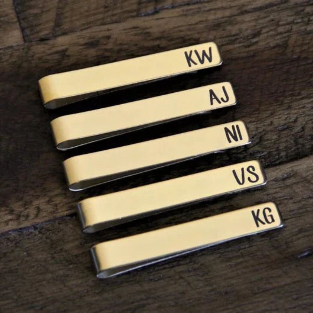 Set of five brass tie bars engraved with sample initials KW, AJ, NI, US, and KG, displayed on dark wood surface