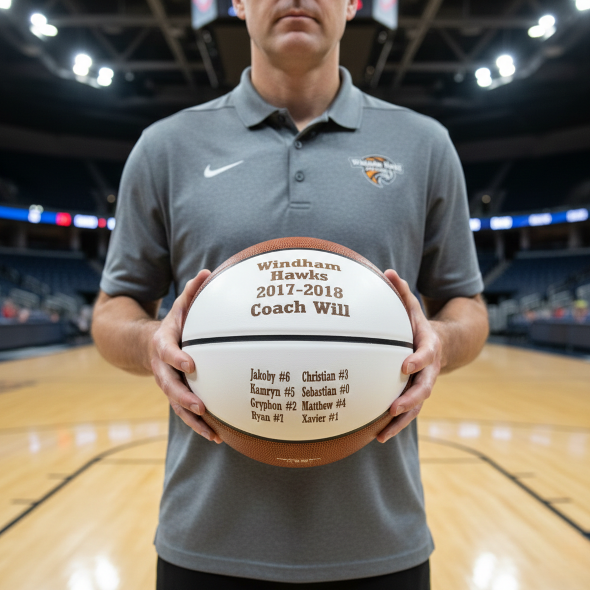 Basketball engraved with sample text "Windham Hawks 2017-2018 Coach Will" and sample roster names, held by coach on indoor court