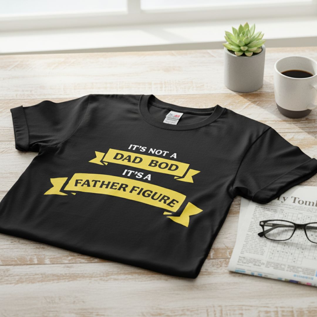 Black crewneck t-shirt with yellow banner design reading "It's not a dad bod it's a father figure" displayed flat on wooden surface with coffee mug and succulent plant